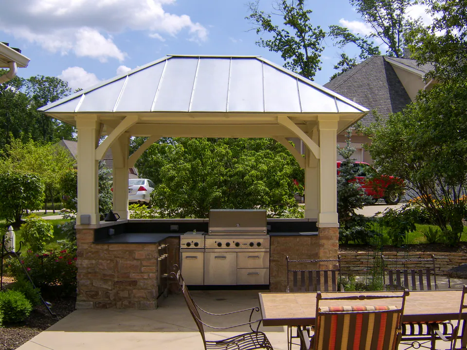 Covered outdoor kitchen pavilion with stainless grill, outdoor cabinets, stone bases, standing seam roof, paver patio, and dining nearby